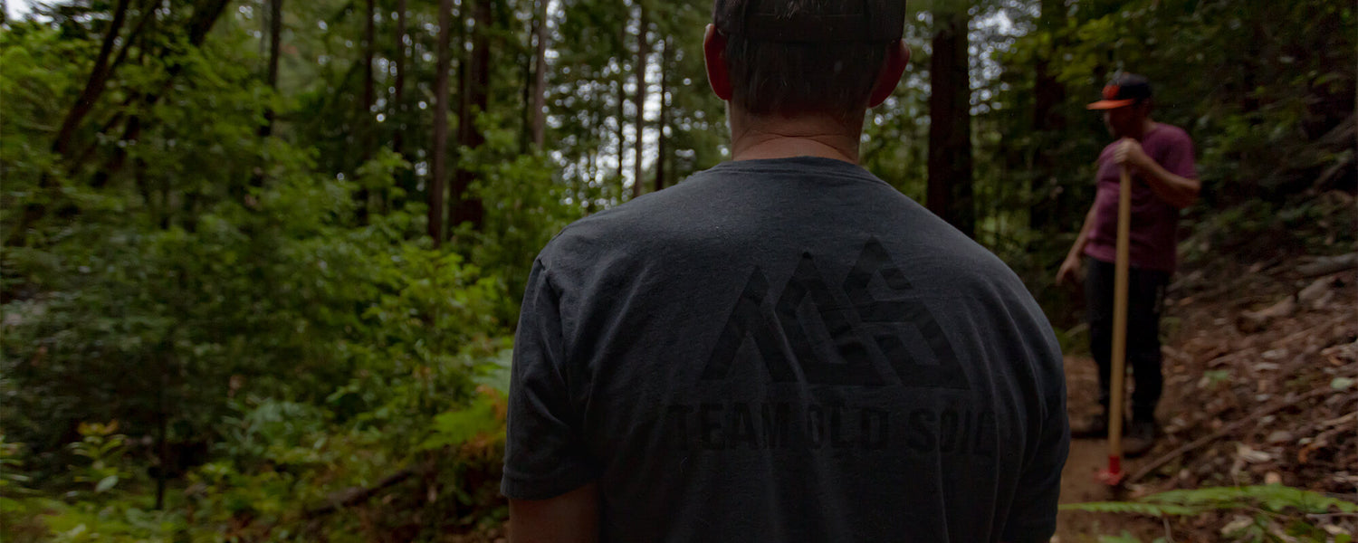 Two men digging in the forest. Wearing charcoal class TOS shirt. Second male is holding a rake wearing Maroon Traction TOS shirt. Local trail builders.